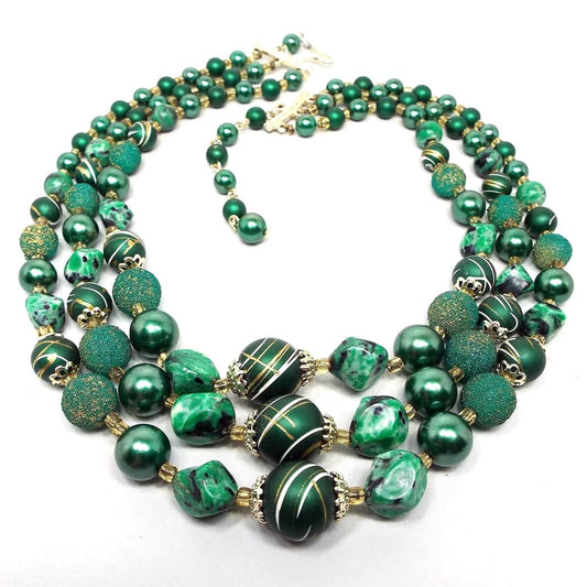 Top view of the Japan Mid Century multi strand beaded necklace. The necklace has three strands of beads. There are pearly round, textured round, paint striped round, and nugget style beads in varying shades of green. There is a hook clasp at the end.