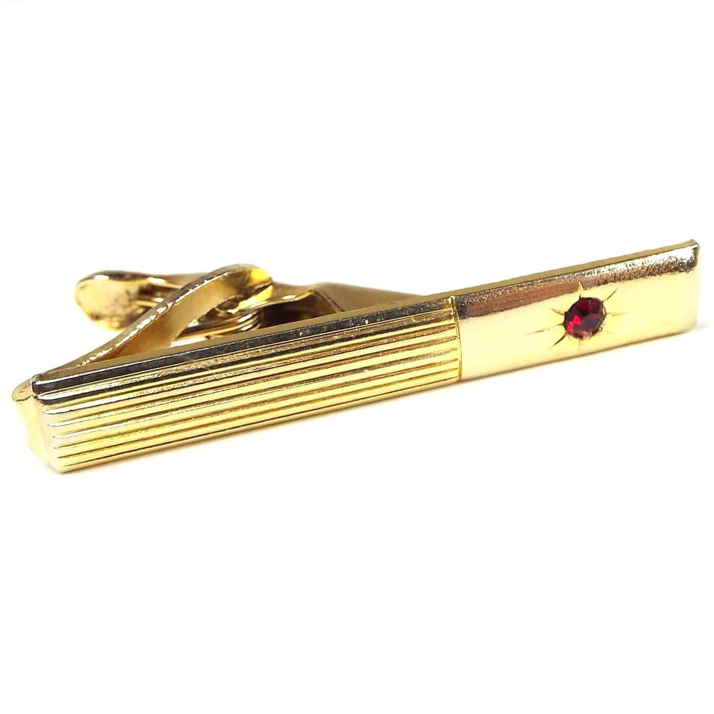 Front view of the retro vintage rhinestone tie clip. The metal is gold tone in color. There are thin raised stripes on one end and the other end has a starburst design with a red rhinestone in the middle.
