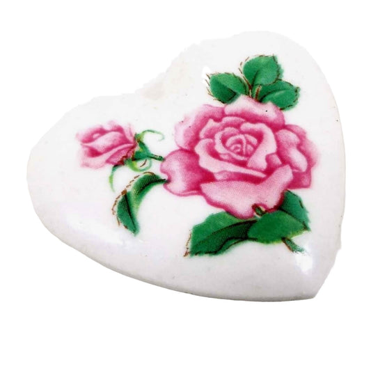 Angled view of the retro vintage Avon ceramic floral heart brooch. The ceramic is white in color. It has a decal on the front of two pink roses with green leaves.