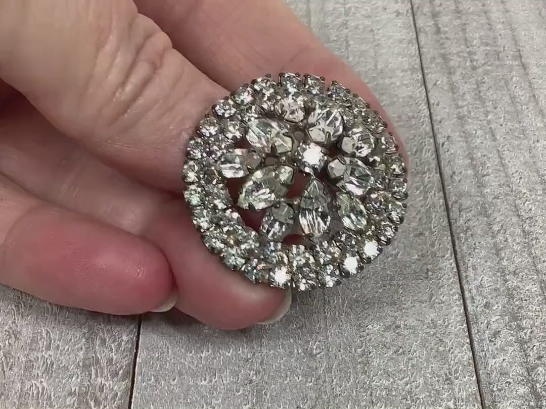 Video of the Mid Century vintage rhinestone brooch pin. It is silver tone in color and round in shape. There are two rows of round rhinestones around the edge and a floral like design of round and marquis rhinestones in the middle. The video shows how they sparkle.