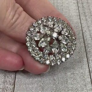 Video of the Mid Century vintage rhinestone brooch pin. It is silver tone in color and round in shape. There are two rows of round rhinestones around the edge and a floral like design of round and marquis rhinestones in the middle. The video shows how they sparkle.