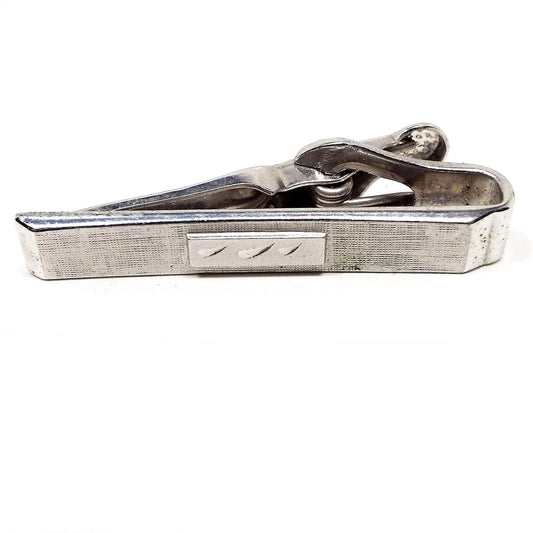 Front view of the Mid Century vintage Swank tie clip. It is silver tone in color and has a matte textured front. The middle has a shiny silver tone rectangle with three etched teardrop shapes in it.