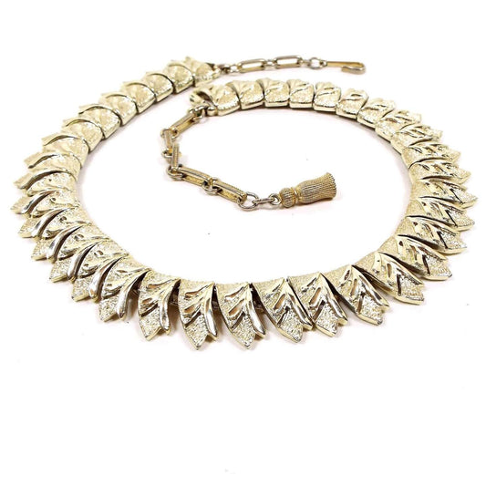 Front view of the Mid Century vintage Coro link choker necklace. It is gold tone in color. The links are shaped like ribbon ends and have an etched leaf type pattern cut on them. There is a hook clasp at the end.