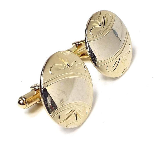 Angled view of the Mid Century vintage gold filled cufflinks. They are round in shape. There is a curved area in the middle with light shiny gold color. On each side of that is matte gold color with an etched leaf like pattern. You can see reflections from items in the room on them.