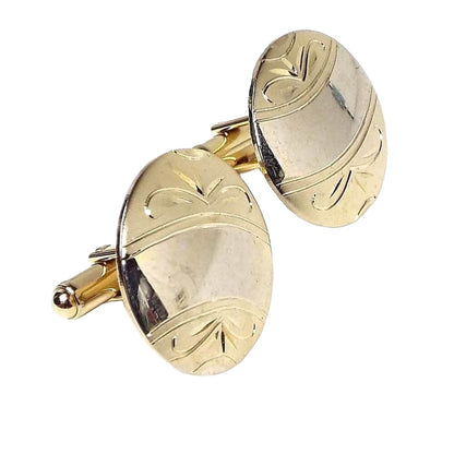 Angled view of the Mid Century vintage gold filled cufflinks. They are round in shape. There is a curved area in the middle with light shiny gold color. On each side of that is matte gold color with an etched leaf like pattern. You can see reflections from items in the room on them.