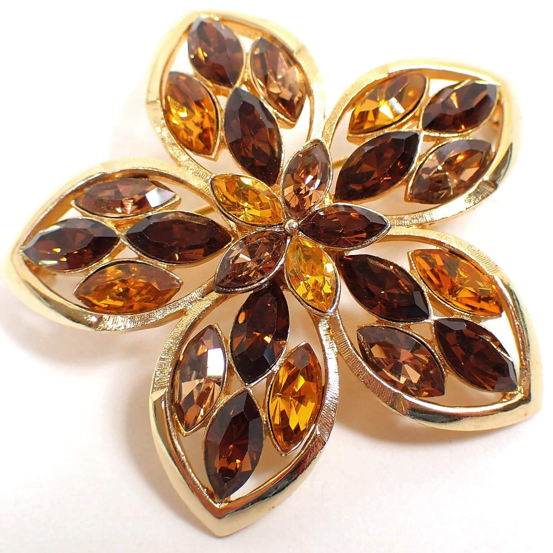 Angled front view of the Mid Century vintage rhinestone floral brooch pin. The metal is gold tone plated in color. There are five rounded petals with pointed ends. Each petal and the middle of the flower has marquis shaped rhinestones in brown, orange, and dark yellow color.