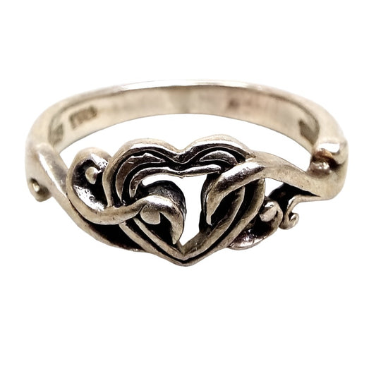 Front view of the retro vintage TMA Thailand sterling silver heart ring. The sterling is slightly darkened from age. The top part of the ring has a heart with open middle design and curls of sterling going over the front into the inside area of the heart giving it a sort of appearance of being held by vines. 