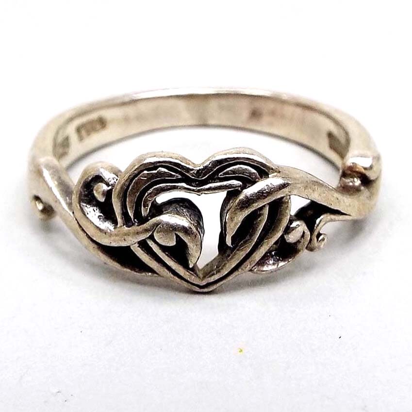 Front view of the retro vintage TMA Thailand sterling silver heart ring. The sterling is slightly darkened from age. The top part of the ring has a heart with open middle design and curls of sterling going over the front into the inside area of the heart giving it a sort of appearance of being held by vines. 