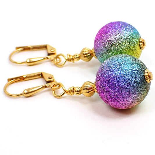 Side view of the handmade metallic rainbow earrings. The metal is gold plated in color. There are large round ball beads at the bottom that are textured and have metallic colors of the rainbow blended all the way around the beads.