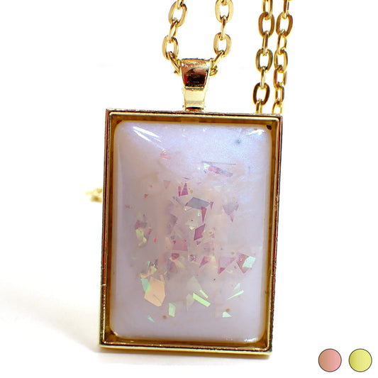 Front view of the large handmade rectangle resin pendant necklace. The resin has an opal like color shift sheen of blue. There is AB pink chunky glitter embedded in the resin. The setting shown is gold plated color but there are two dots at the bottom right of the photo showing a choice of rose gold plated or gold plated.