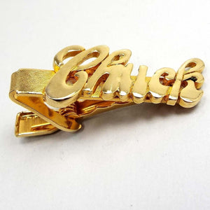 Angled front view of the retro vintage name tie clip. The metal is gold tone in color. The name is shiny and the background is textured. It has the name Chuck in script lettering. There is a small dark spot on the inside area by the K. 