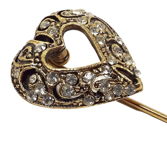 Enlarged view of the retro vintage rhinestone heart stick pin. The metal is antiqued gold tone in color. The heart has an open cut out middle and the rest is filigree and encrusted with clear rhinestones of various sizes. 