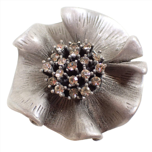 Front view of the retro vintage pewter brooch pendant. It is made of pewter in a medium gray color and is shaped like a rounded ruffle like flower. The middle part of the flower has clear round glass crystal rhinestones. 