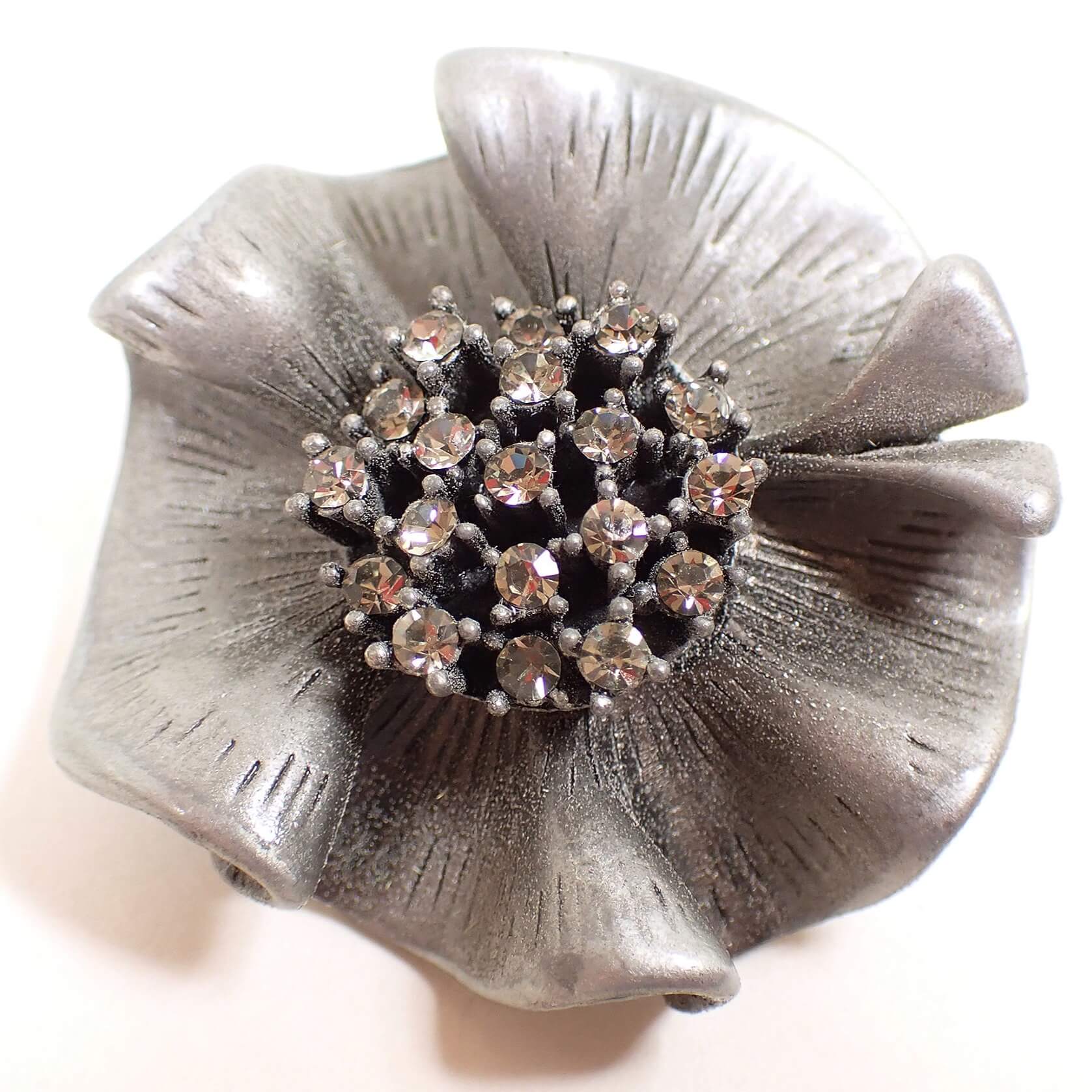 Front view of the retro vintage pewter brooch pendant. It is made of pewter in a medium gray color and is shaped like a rounded ruffle like flower. The middle part of the flower has clear round glass crystal rhinestones. 