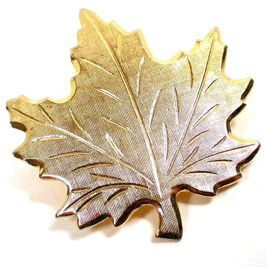 Enlarged front view of the retro vintage leaf pin. The metal is gold tone plated in color. The brooch is shaped like a maple leaf with textured matte metal design and etched leaf veins.