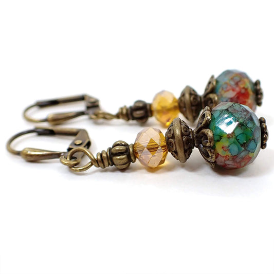 Side view of the handmade faceted glass drop earrings. The metal is antiqued brass in color. There are faceted glass crystals in an orange amber color on the top. The bottom faceted glass beads are multi color with spots of yellow, red, green, and blue, and differ slightly from one another in color pattern.