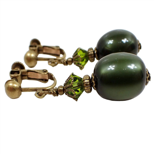 Side view of the handmade olive green oval drop earrings. The metal is antiqued brass in color. There is a faceted glass crystal olive green bead at the top and an oval moonglow lucite olive green bead at the bottom. The lucite bead has a glowy like appearance as you move around in the light.