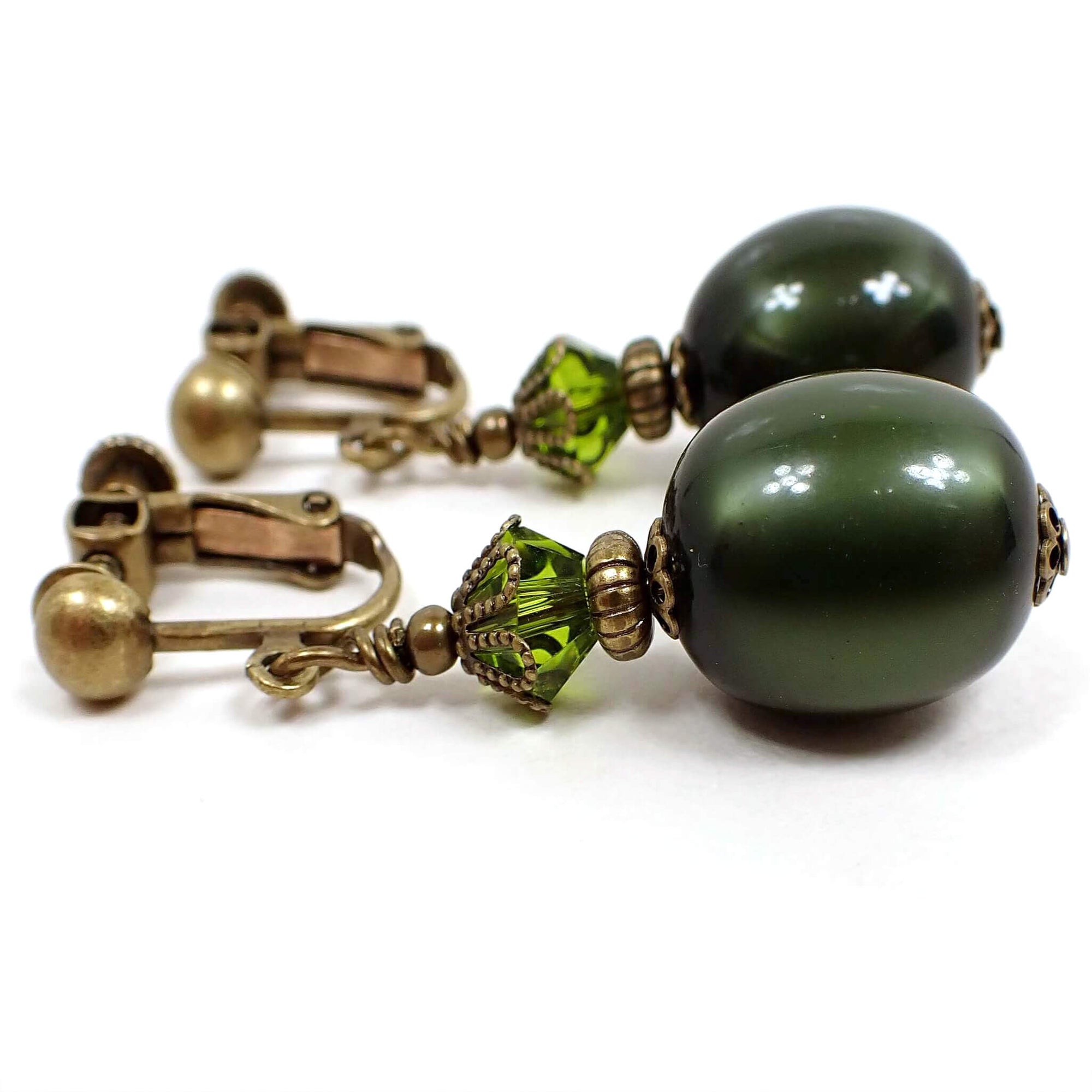 Side view of the handmade olive green oval drop earrings. The metal is antiqued brass in color. There is a faceted glass crystal olive green bead at the top and an oval moonglow lucite olive green bead at the bottom. The lucite bead has a glowy like appearance as you move around in the light.