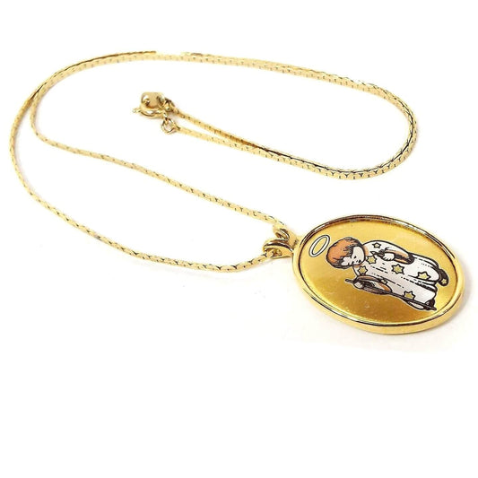 Front view of the Reed and Barton retro vintage pendant necklace. It has a thin gold tone chain with an oval pendant at the bottom. The pendant is Damascene style with a depiction of a child with a halo, a robe with stars on it, and a staff. The metal is gold tone in color and there is copper color and white on the pendant as well.