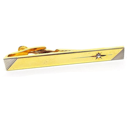 Front view of the retro vintage two tone tie clip. It is mostly gold tone in color with silver tone etched diagonal lines on the ends. There is a starburst cut design on one end with a rhinestone in the middle.