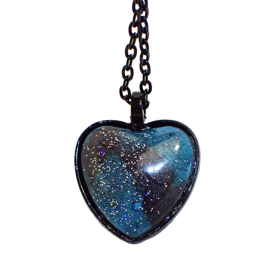 Enlarged front view of the Goth heart pendant necklace with handmade resin cab. The chain and pendant setting are black coated. The resin on the heart pendant has pearly teal blue and dark gray resin with tiny specks of holographic glitter for bits of sparkle and flashes of color.