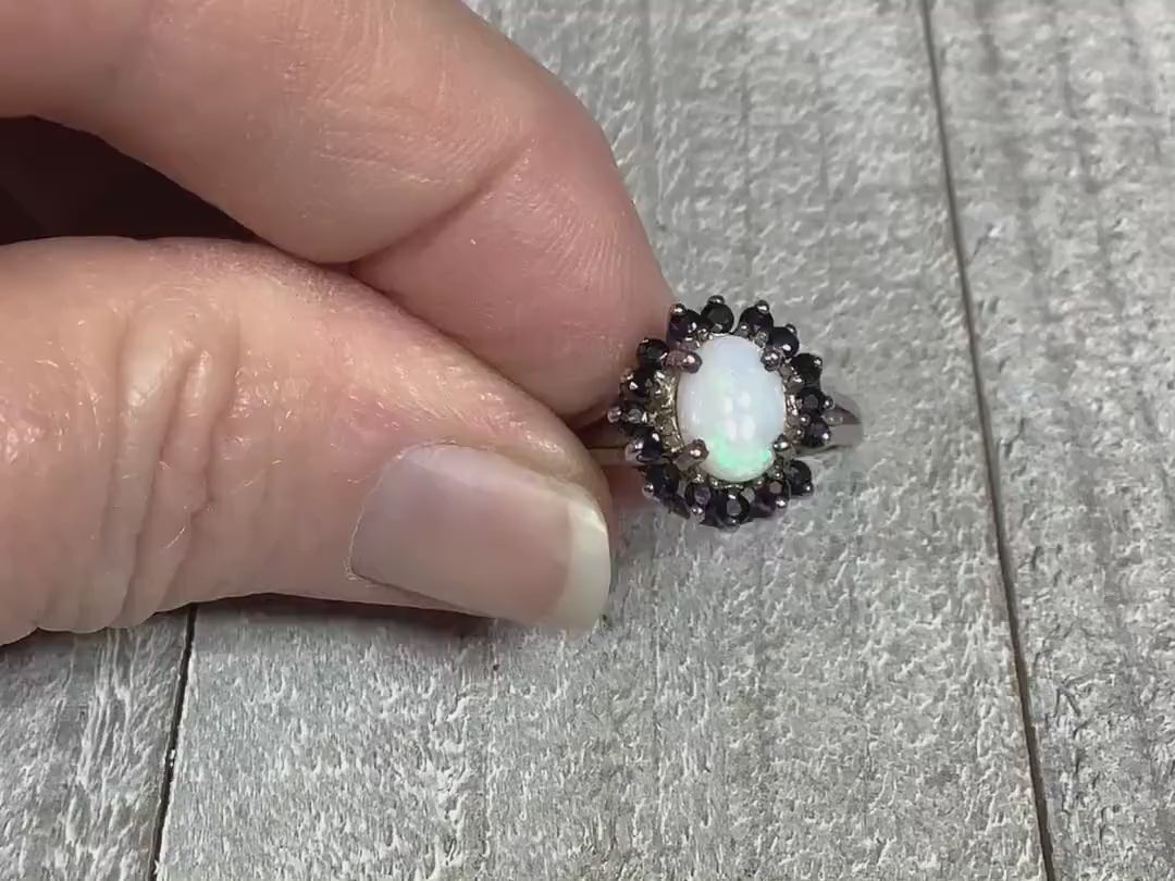 Video of the retro vintage sterling silver ring with small sapphires and simulated opal. The simulated opal is oval shaped and in the middle of the ring. It is surrounded by small round blue sapphire gemstones. The video shows the flash and sparkle of the ring.