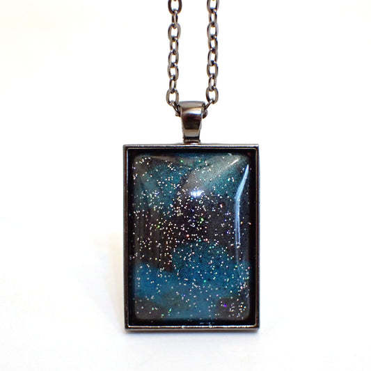 Front view of the large rectangle handmade resin pendant necklace. The metal is gunmetal dark gray in color. The resin cab on the front has marbled areas of pearly dark gray and teal blue resin. There is tiny specks of silver holographic glitter scattered around the cab for bits of sparkle and flashes of color.