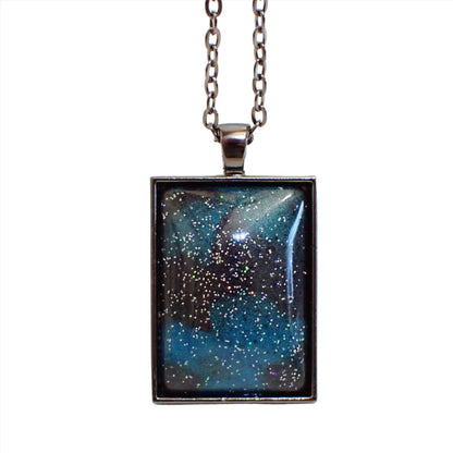 Front view of the large rectangle handmade resin pendant necklace. The metal is gunmetal dark gray in color. The resin cab on the front has marbled areas of pearly dark gray and teal blue resin. There is tiny specks of silver holographic glitter scattered around the cab for bits of sparkle and flashes of color.