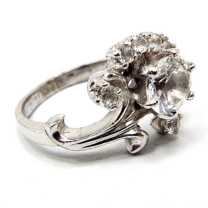 Angled front and side view of the Mid Century vintage sterling silver Quartz ring. The top part has a flower design with curved stem and leaves. There is a prong set round quartz gemstone in the middle of the flower and smaller sized prong set round quartz gemstones around the top of it. 