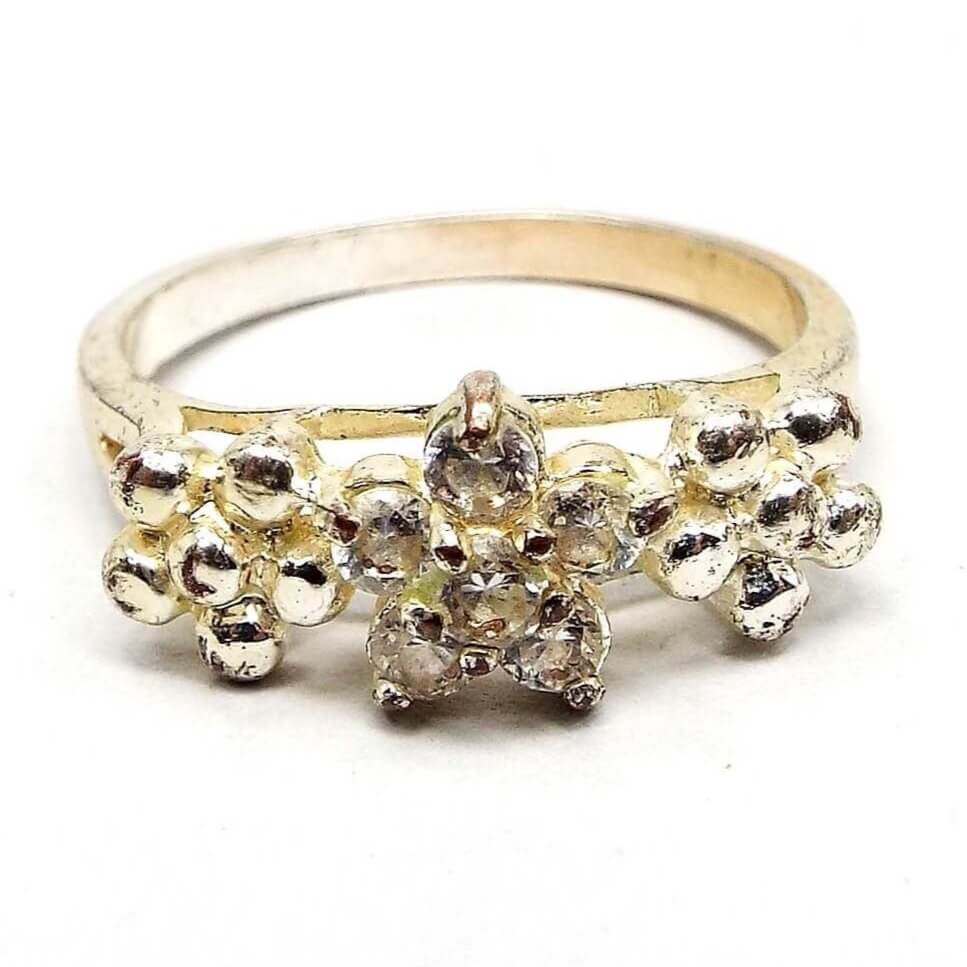 Front view of the retro vintage rhinestone floral ring. The metal is silver tone in color, but has a slight yellowing from age. There are three flowers on the top part of the ring over a split band. The middle flower has five prong set round clear rhinestones. The other two flowers are made of round metal balls and are on each side of the rhinestone flower.