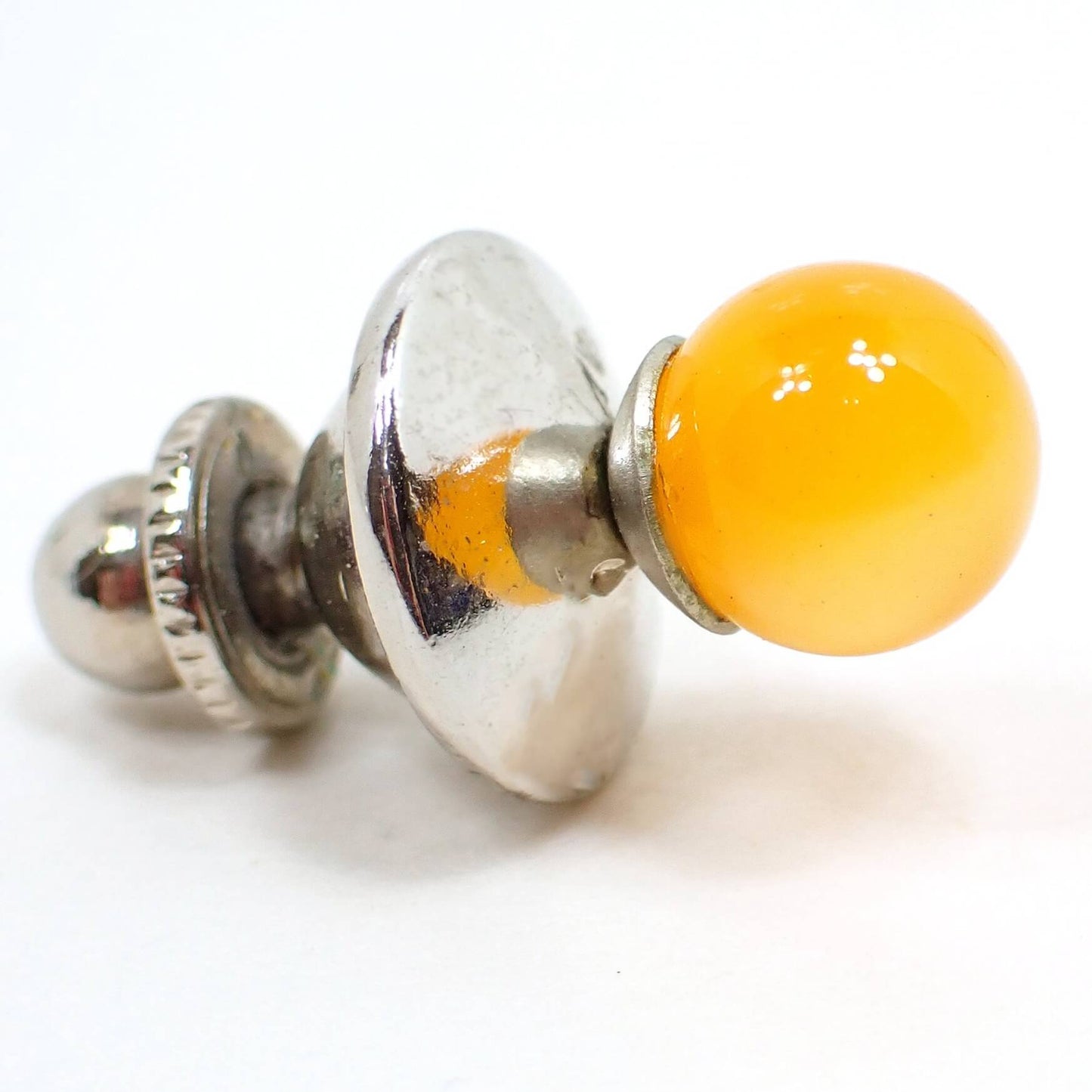 Angled side view of the Mid Century vintage moonglow lucite stud tie tack. The metal is silver tone plated in color. There is a round orange lucite sphere on the front. Moonglow lucite has an inner glow like appearance as you move around in the light.
