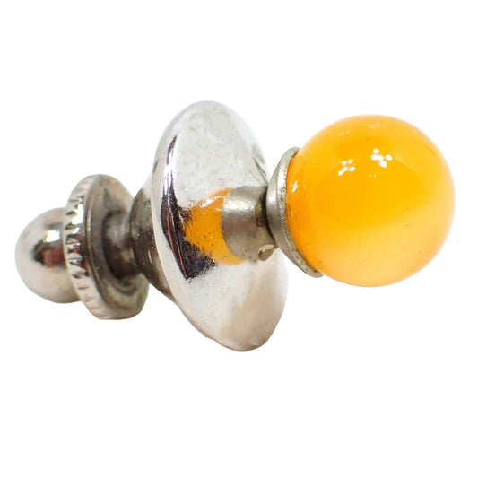 Angled side view of the Mid Century vintage moonglow lucite stud tie tack. The metal is silver tone plated in color. There is a round orange lucite sphere on the front. Moonglow lucite has an inner glow like appearance as you move around in the light.