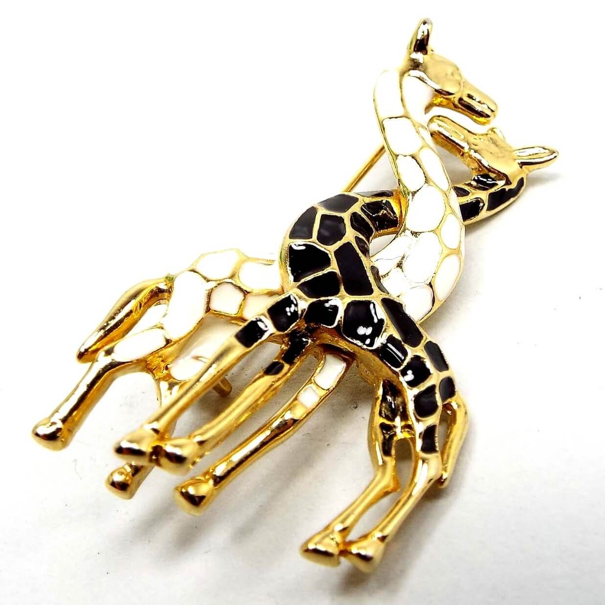 Front view of the retro vintage giraffe brooch. the metal is gold tone in color. There are two giraffes with their necks intertwined. One has white enameled spots and the other has black enameled spots.
