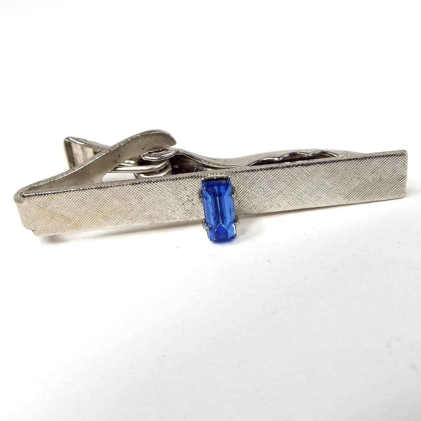 Front view of the retro vintage blue rhinestone tie clip. The metal is silver tone in color and has a brushed textured design. In the middle is a prong set baguette rhinestone in blue. 