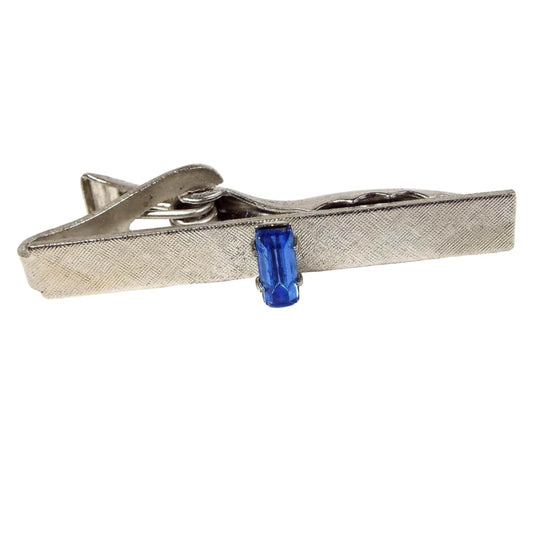 Front view of the retro vintage blue rhinestone tie clip. The metal is silver tone in color and has a brushed textured design. In the middle is a prong set baguette rhinestone in blue. 