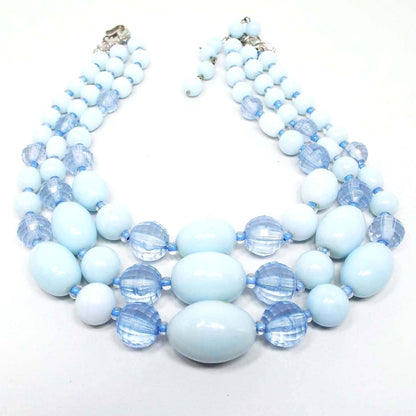 Front view of the Mid Century vintage multi strand beaded necklace. There are three strands of larger sized chunky beads. The beads are round, faceted round, and oval shaped and are graduated in size with the largest at the bottom of the necklace. The faceted beads are blue acrylic and the others are light blue smooth lucite. 