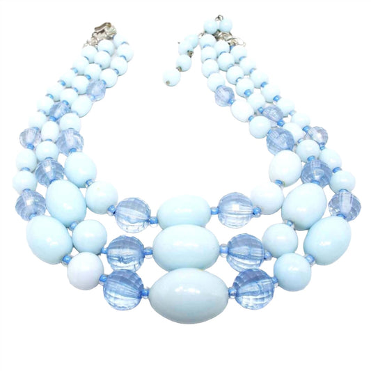 Front view of the Mid Century vintage multi strand beaded necklace. There are three strands of larger sized chunky beads. The beads are round, faceted round, and oval shaped and are graduated in size with the largest at the bottom of the necklace. The faceted beads are blue acrylic and the others are light blue smooth lucite. 