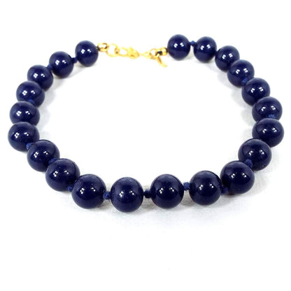 Angled view of the retro vintage Monet beaded bracelet. The beads are round and dark blue in color. There are knots tied between each bead and a hinged clip clasp in gold tone metal at the end.