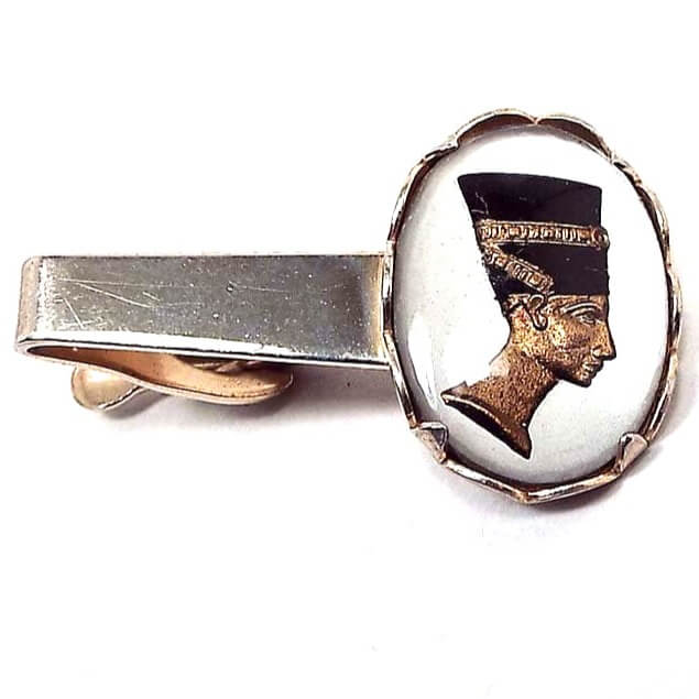 Front view of the Mid Century vintage Egyptian revival tie clip. It is gold tone in color and has a large oval on the end. In the oval is a bezel and prong set glass cab with an intaglio cameo of Nefertiti in black, white, and gold color. 