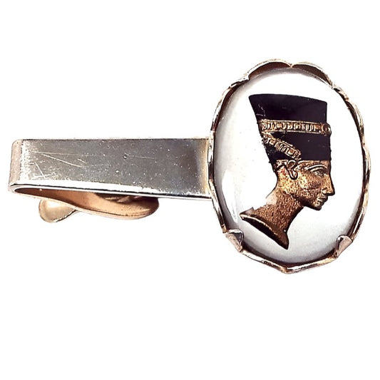 Front view of the Mid Century vintage Egyptian revival tie clip. It is gold tone in color and has a large oval on the end. In the oval is a bezel and prong set glass cab with an intaglio cameo of Nefertiti in black, white, and gold color. 
