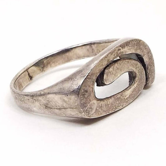Angled front and side view of the retro vintage sterling silver Modernist style swirl ring. The sterling is darkened from age. There is a dark line inside the bottom band area. The top has a band of sterling that curves around the top and then curls inward at the bottom. 