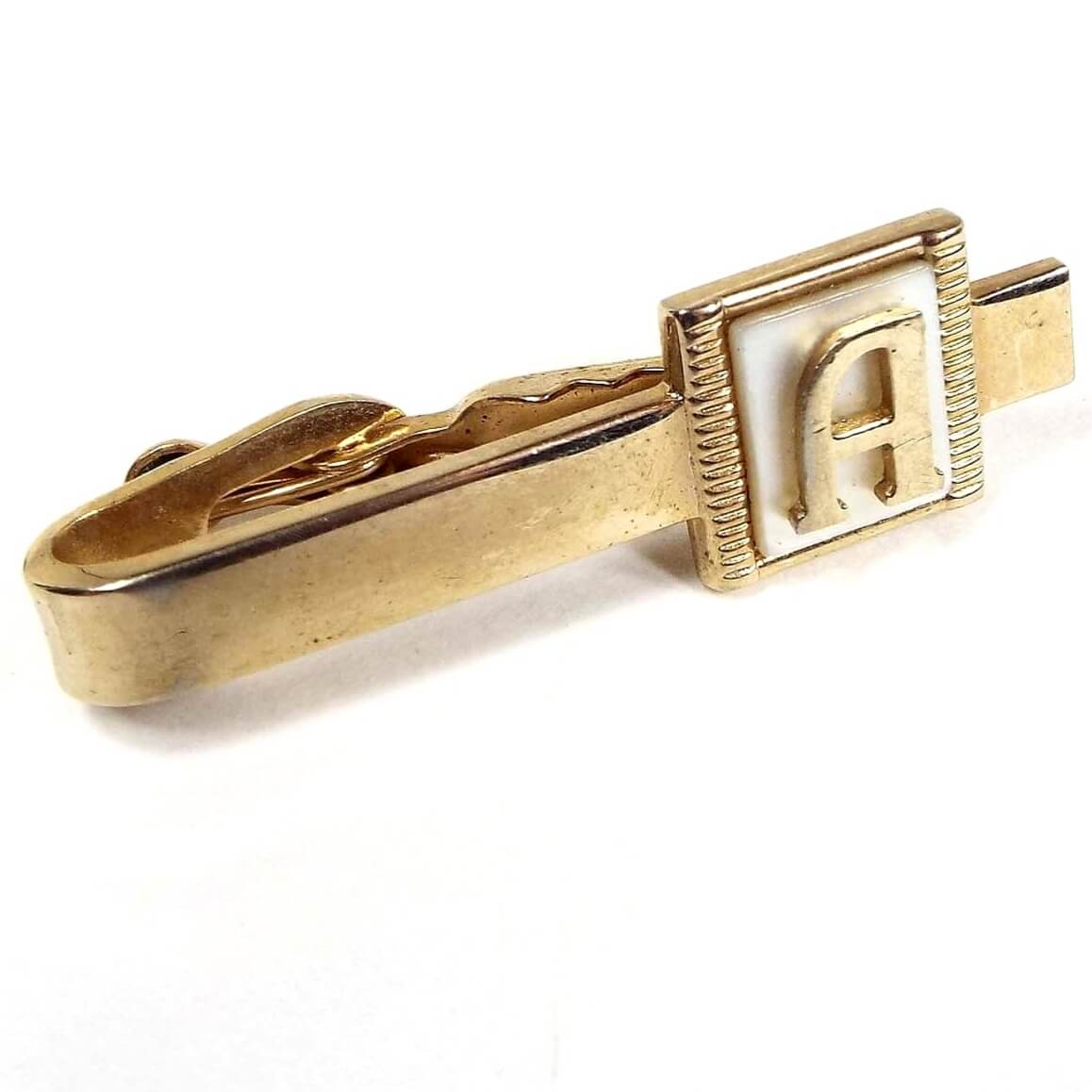 Angled front view of the Mid Century vintage initial tie clip. It is gold tone in color and has a square pearly white mother of pearl shell cab on the end. There is a letter A on the front. 
