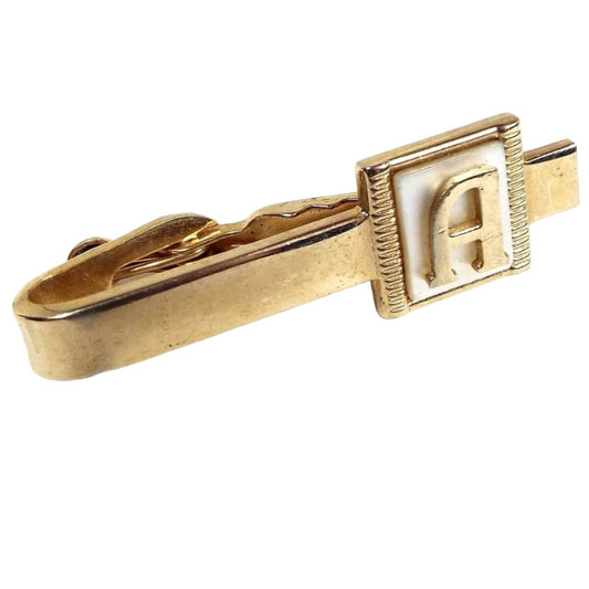Angled front view of the Mid Century vintage initial tie clip. It is gold tone in color and has a square pearly white mother of pearl shell cab on the end. There is a letter A on the front. 