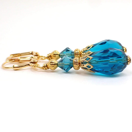 Side view of the handmade teardrop earrings. The metal is gold plated in color. There are faceted glass crystal rondelle beads at the top and faceted glass crystal teardrop beads at the bottom. The beads are teal blue in color.