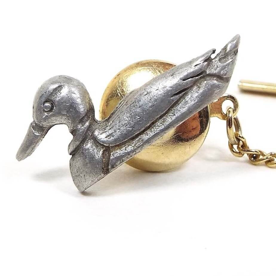 Enlarged front view of the retro vintage pewter tie tack. It is shaped like a 3D duck in the water with a flat bottom. It is gray in color and the clutch back and chain is gold tone in color.