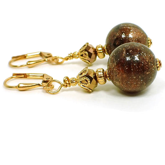 Angled side view of the handmade sparkly brown lucite galaxy earrings. The metal is gold plated in color. It has a faceted glass metallic brown bead at the top and a round lucite bead at the bottom that has tiny sparkles and swirls of brown and black that resembles a galaxy like appearance.