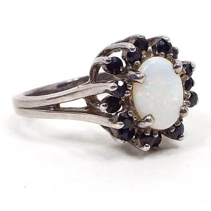 Angled front and side view of the retro vintage sterling silver sapphire and faux opal ring. The band is split style at the top. There is an oval simulated opal in the middle with small round sapphire gemstones in varying shades of dark blue all the way around the faux opal. 