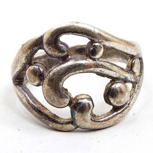 Front view of the retro vintage sterling silver band ring. The top part of the ring has a wide cut out style area with curls and round dots. The sterling is slightly darkened from age. The ring tapers on the sides with the widest part being in the middle of the top part of the ring.