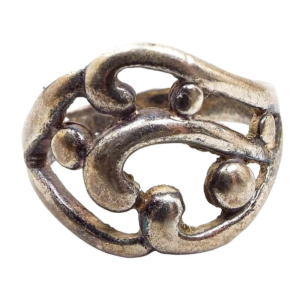 Front view of the retro vintage sterling silver band ring. The top part of the ring has a wide cut out style area with curls and round dots. The sterling is slightly darkened from age. The ring tapers on the sides with the widest part being in the middle of the top part of the ring.