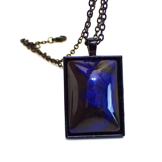 Enlarged front view of the Goth handmade resin pendant necklace. The metal is black coated, including the chain and lobster clasp. There is a large rectangle pendant with a domed resin cab on the front. The handmade resin cab has marbled areas of pearly black and cobalt blue resin.